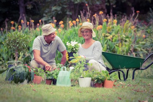 A cheerful elderly man and woman sitting in a well-maintained garden with a lush green lawn, surrounded by various potted plants and flowering shrubs; the woman is holding a blooming plant while the man appears to be engaging in conversation, both wearing wide-brimmed hats for sun protection. Behind them, a vibrant flower border with tall purple and orange flowers adds colour to the garden, which is enclosed by leafy trees. A garden watering can is placed among the potted plants, and a green wheelbarrow is positioned nearby, suggesting ongoing gardening activities. The scene is set outdoors on a bright, clear day, with natural sunlight illuminating the garden and highlighting the different textures of grass, soil, and plant foliage. This image reveals a peaceful and well-organised outdoor space suitable for gardening services in Morden, with attention to plant care and garden aesthetics, emphasizing the importance of lawn and plant maintenance for a thriving garden environment.