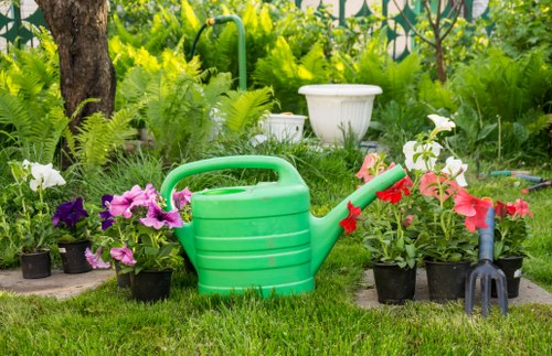 In a well-maintained garden setting, there is a collection of potted flowering plants with vibrant pink, purple, and white blossoms arranged along the edge of a lush, green lawn. Behind the plants, a white decorative stone planter is partially visible among dense greenery, including various shrubs and a tree trunk on the left side of the image. In the foreground, a green plastic watering can with a red spout rests on the grass, surrounded by gardening tools such as a small black hand rake with a red handle and additional potted plants. The background features a mix of leafy foliage and garden features that suggest a neatly tended outdoor space in Morden, ideal for gardening and lawn care services. The garden appears to be taken on a bright, sunny day, with natural daylight illuminating the vibrant plant colours and healthy grass, enhancing the overall impression of a cared-for residential garden that benefits from professional gardening services to maintain its tidy appearance.