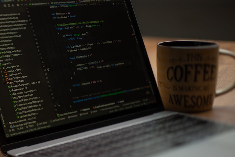A close-up of a laptop displaying lines of code on a dark-themed programming interface, with a wooden table surface underlining the scene. To the right of the laptop, there is a ceramic mug with a bei
